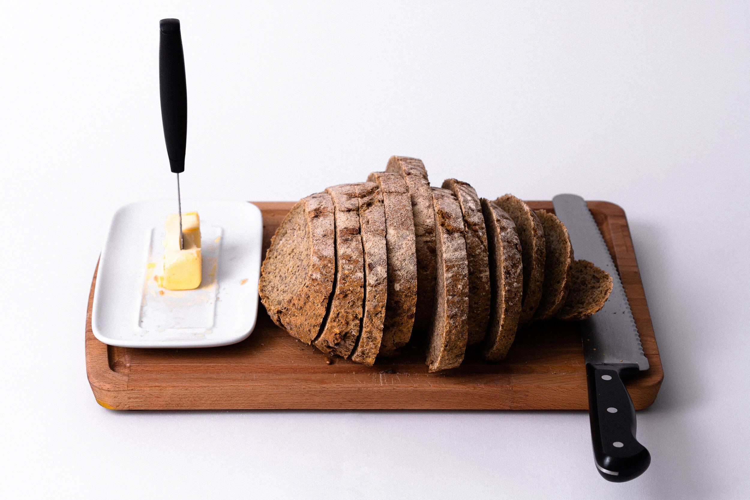 Sliced whole grain bread on a wooden cutting board, accompanied by a butter dish and a butter knife, showcasing a rustic and inviting kitchen setting.