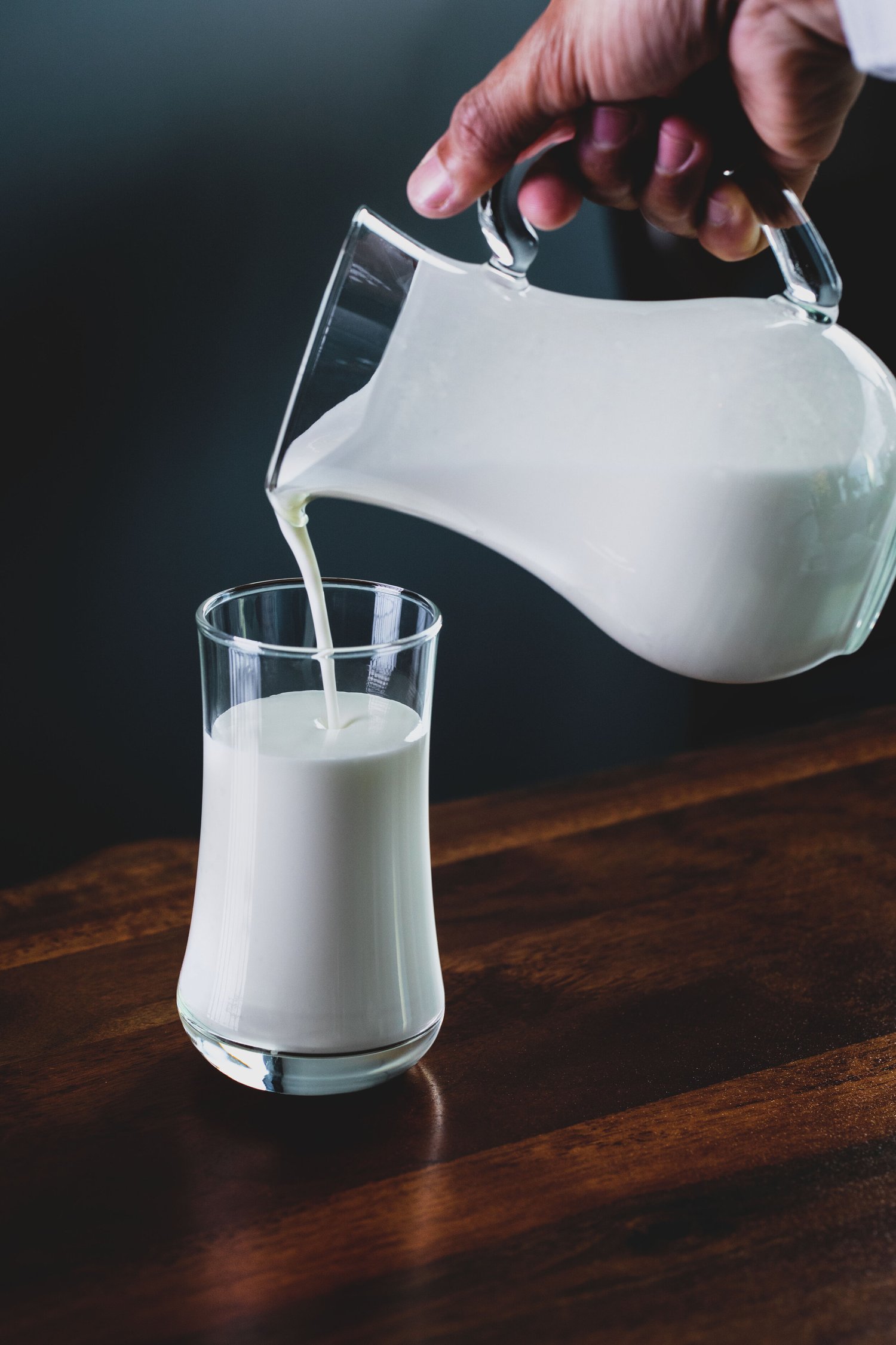 A hand pouring fresh milk from a glass pitcher into a tall, clear glass on a wooden table, showcasing the creamy texture and color of the milk against a dark background.