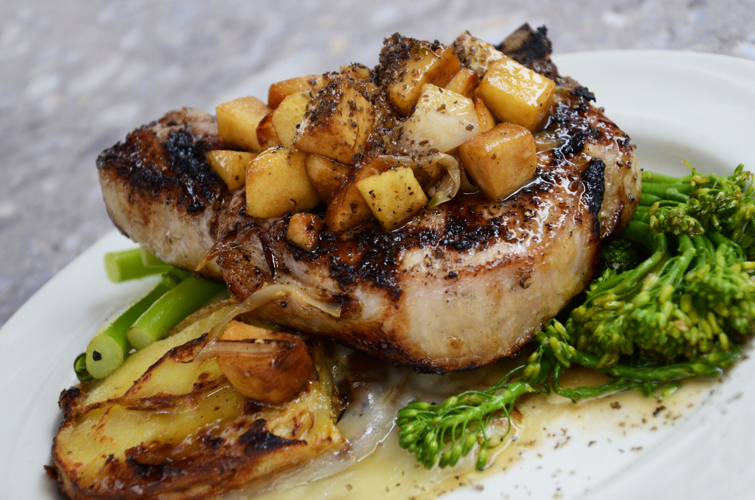 Grilled pork chop topped with sautéed apples and onions, served with roasted potatoes and steamed broccolini on a white plate.