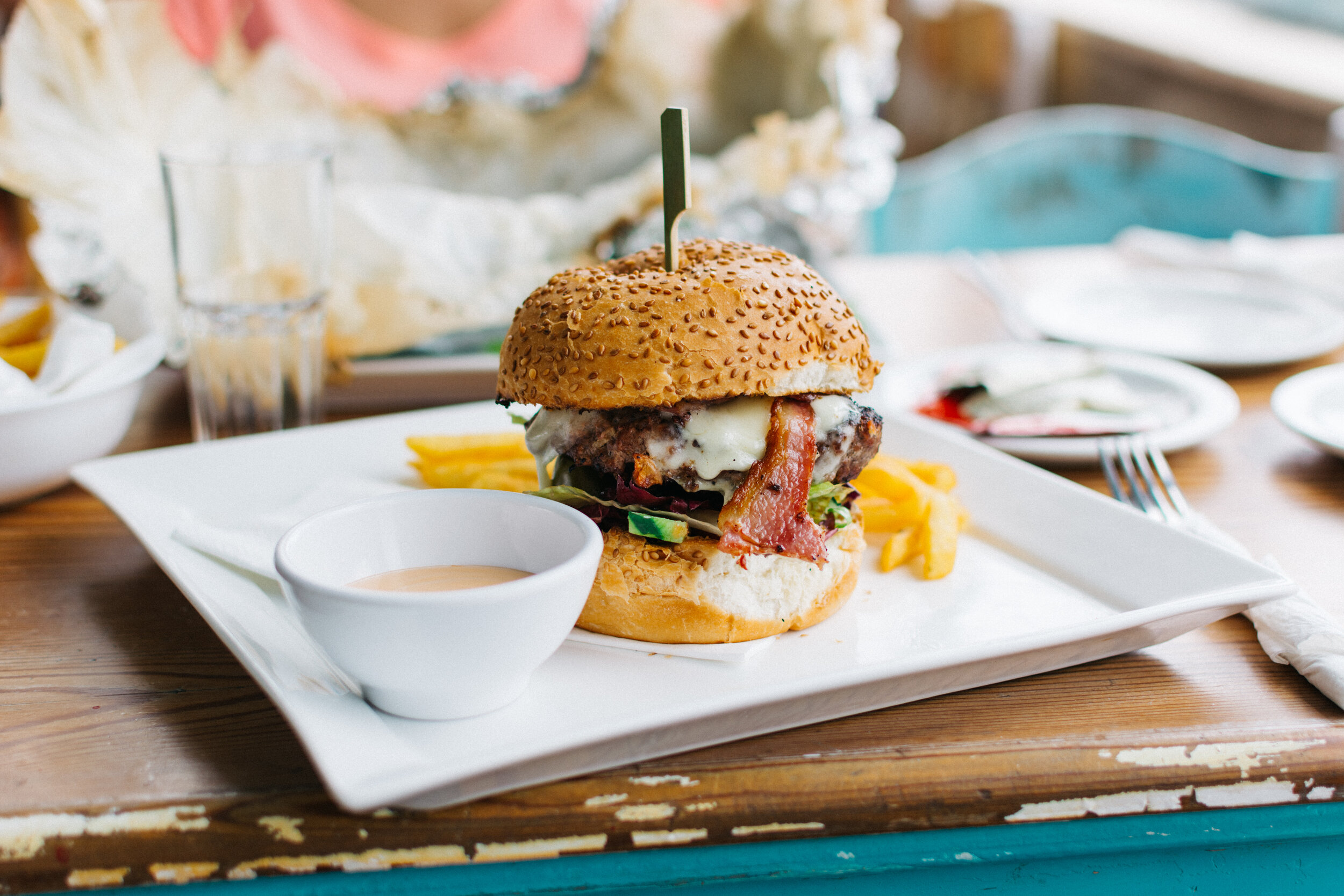 Delicious cheeseburger topped with crispy bacon, fresh lettuce, and melted cheese, served with golden French fries and a side of dipping sauce, on a white plate in a casual dining setting.