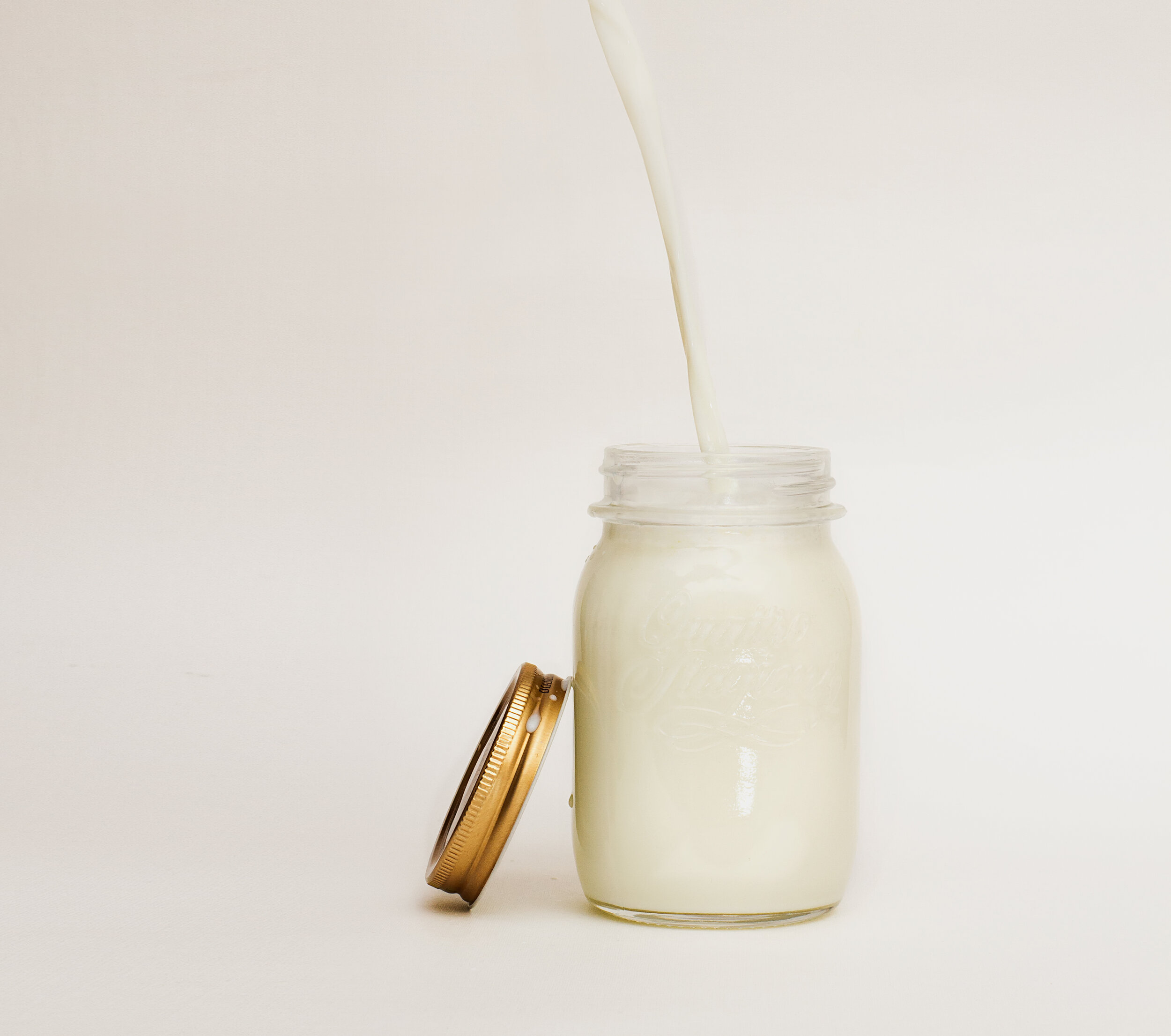 Fresh milk being poured into a clear glass jar with a golden lid, showcasing a creamy texture against a light background. Ideal for recipes or dairy product promotions.