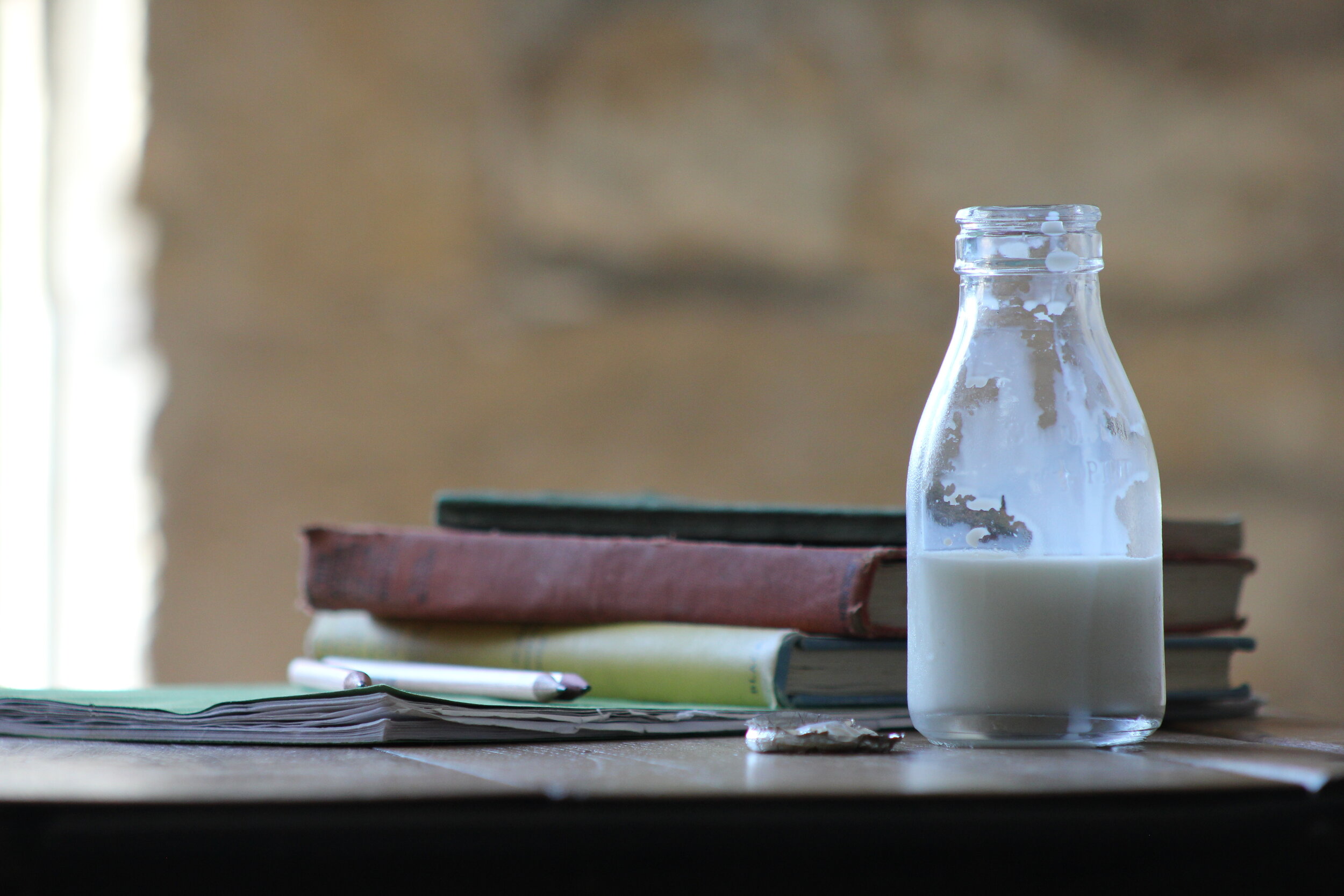 A vintage glass bottle of milk sits on a wooden table next to a stack of old books and a pen, with a blurred background creating a cozy study atmosphere.
